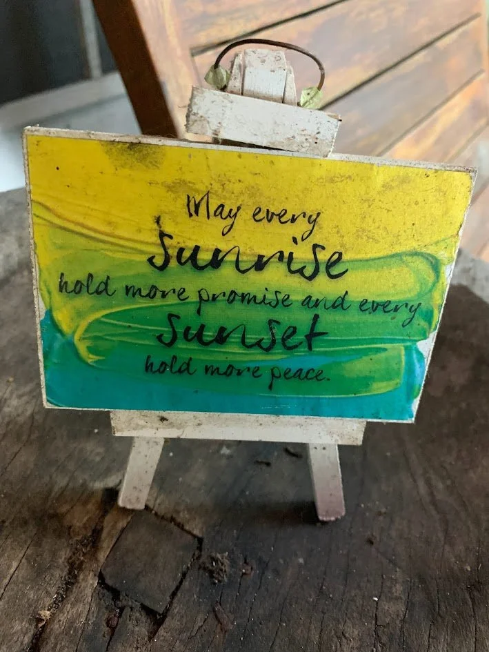 Small wooden sign featuring a colorful watercolor background with yellow, green, and blue swirls. The sign has a handwritten quote: "May every sunrise hold more promise and every sunset hold more peace." It is placed on a miniature easel on a dark wo