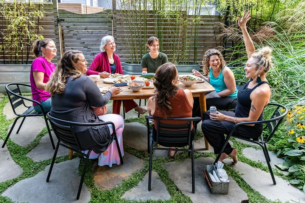 A group of seven women enjoying a meal together outdoors, sitting around a wooden table in a lush garden setting.