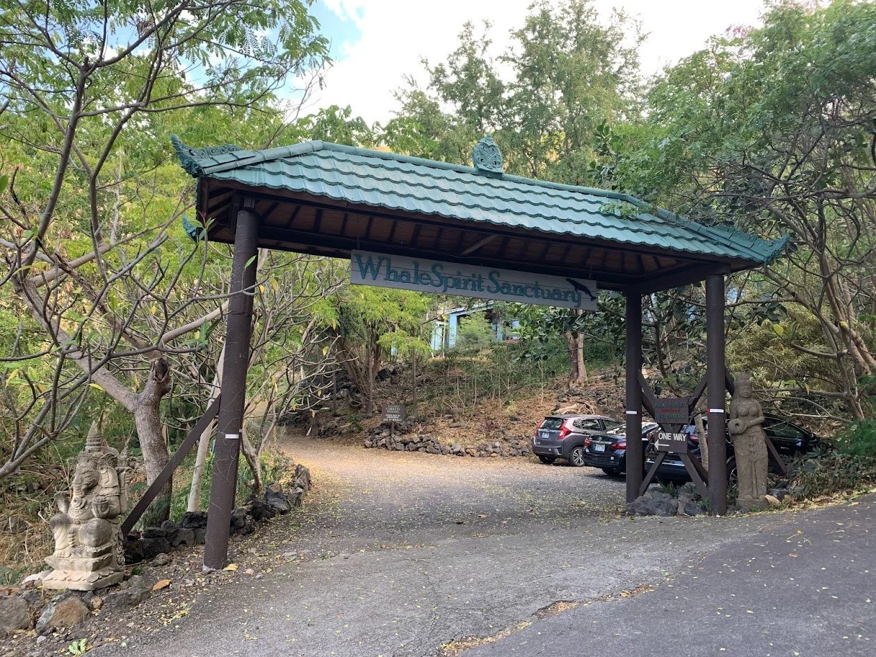Entrance to Whale Spirit Sanctuary with a traditional-style roof, surrounded by trees, cars parked nearby, and stone statues on each side of the entrance