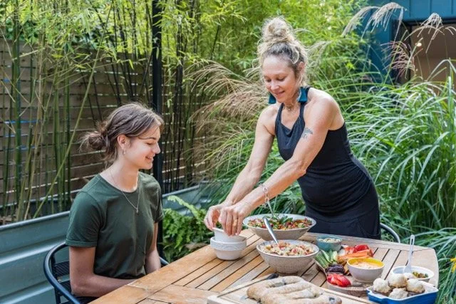 A woman preparing food on a wooden outdoor table surrounded by greenery, while a girl sits nearby, smiling.