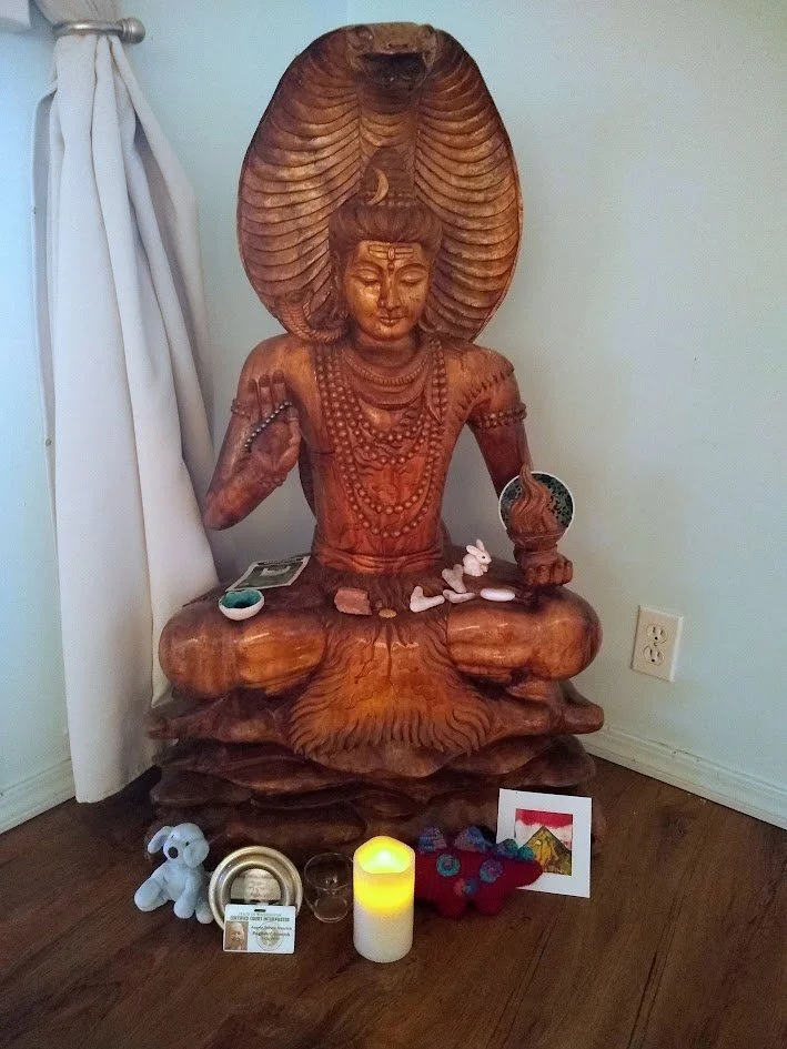 Wooden carved statue of a seated figure with a large, elaborate headdress, surrounded by small offerings and candles on the floor.