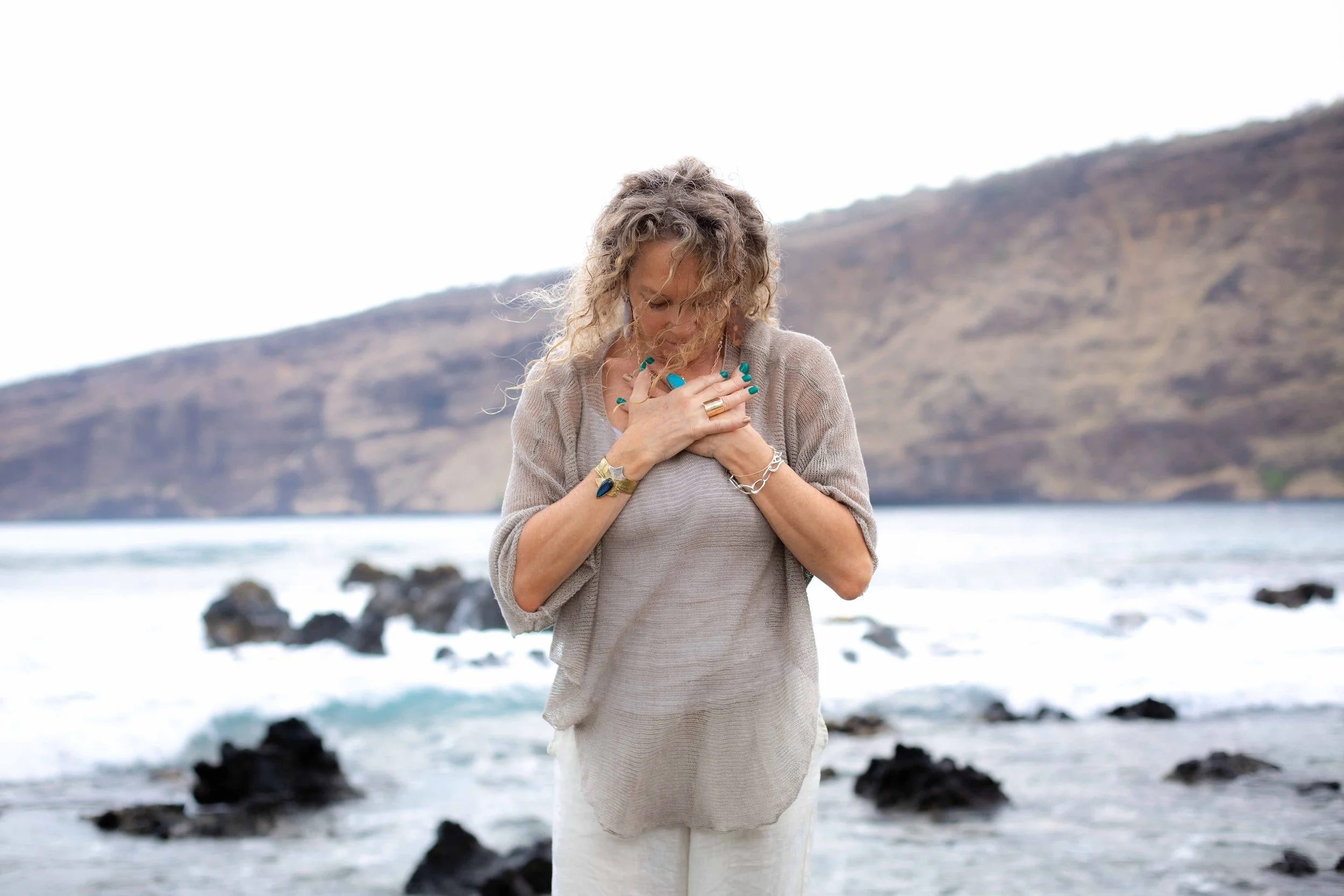 A woman with curly hair wearing a beige cardigan and white dress, standing on a beach with rocks and mountains in the background, looking down with her hands clasped over her chest.