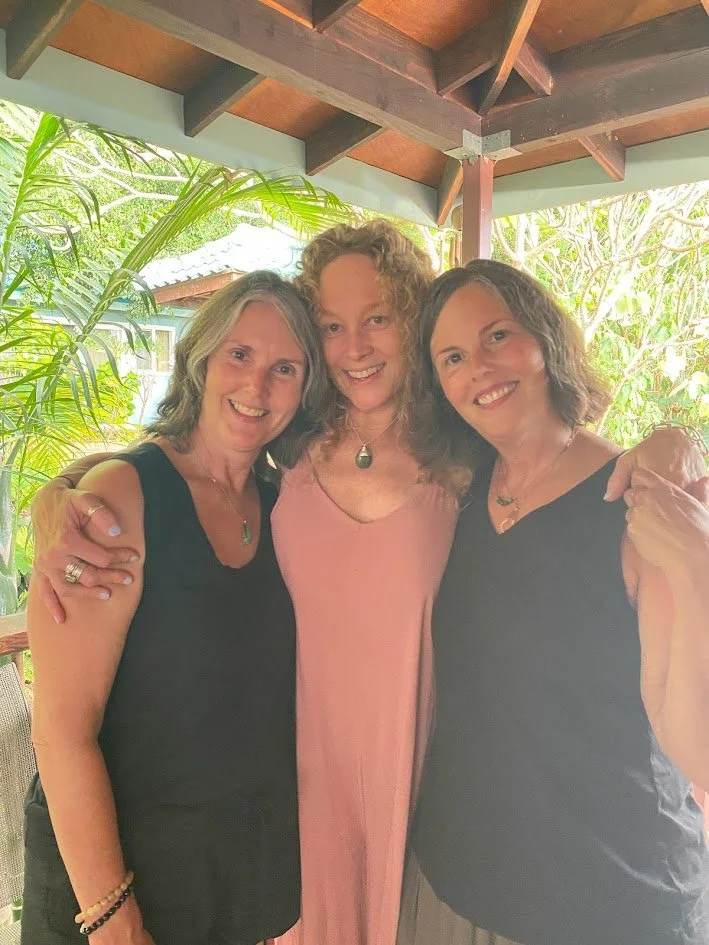 Three women smiling and standing close together, outdoors under a wooden roof with green foliage in the background.