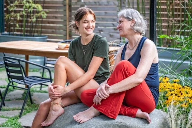 Two women sitting on a large stone in a backyard garden, smiling and engaging in a conversation, with flowers and outdoor furniture in the background.