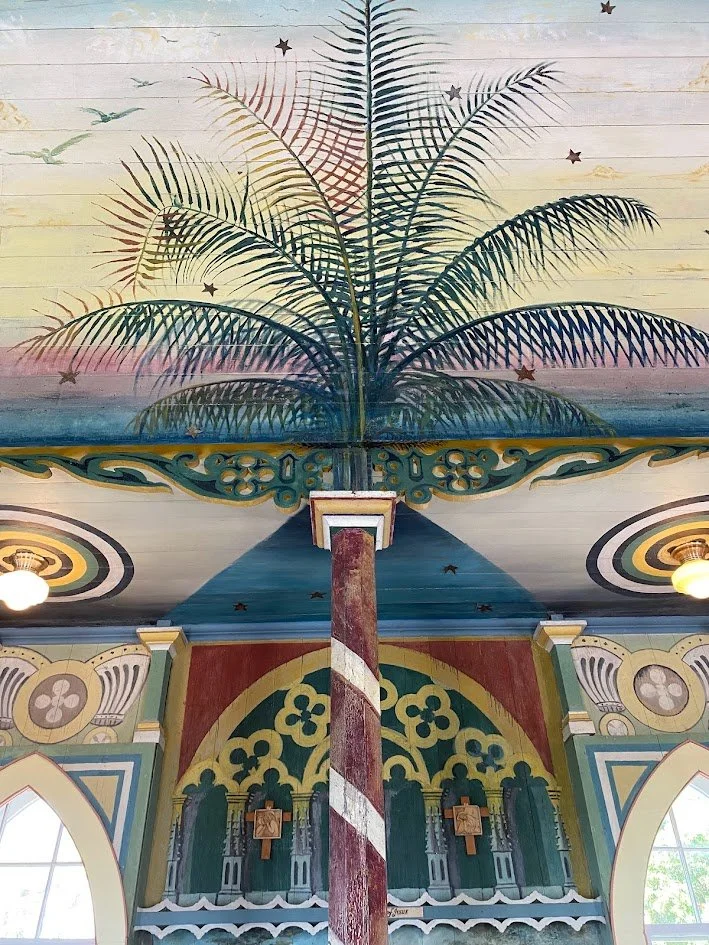 Decorative ceiling featuring a painted palm tree, stars, and a sunset sky, supported by a striped red and white column, inside a colorful building with arched windows and ornate designs.