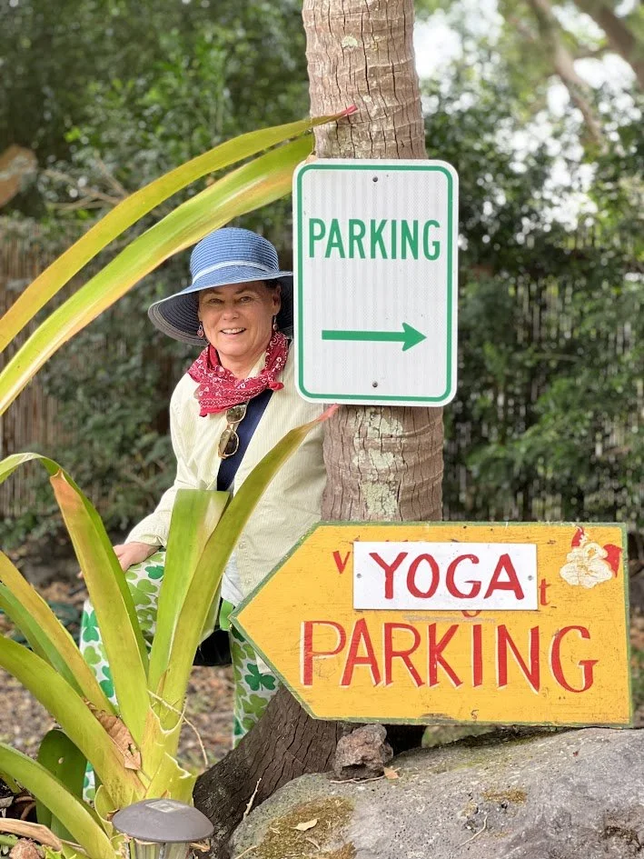 A woman wearing a wide-brimmed blue hat, red bandana, cream shirt, and green patterned pants standing behind a tree with signs pointing to parking and yoga parking in a lush outdoor setting.