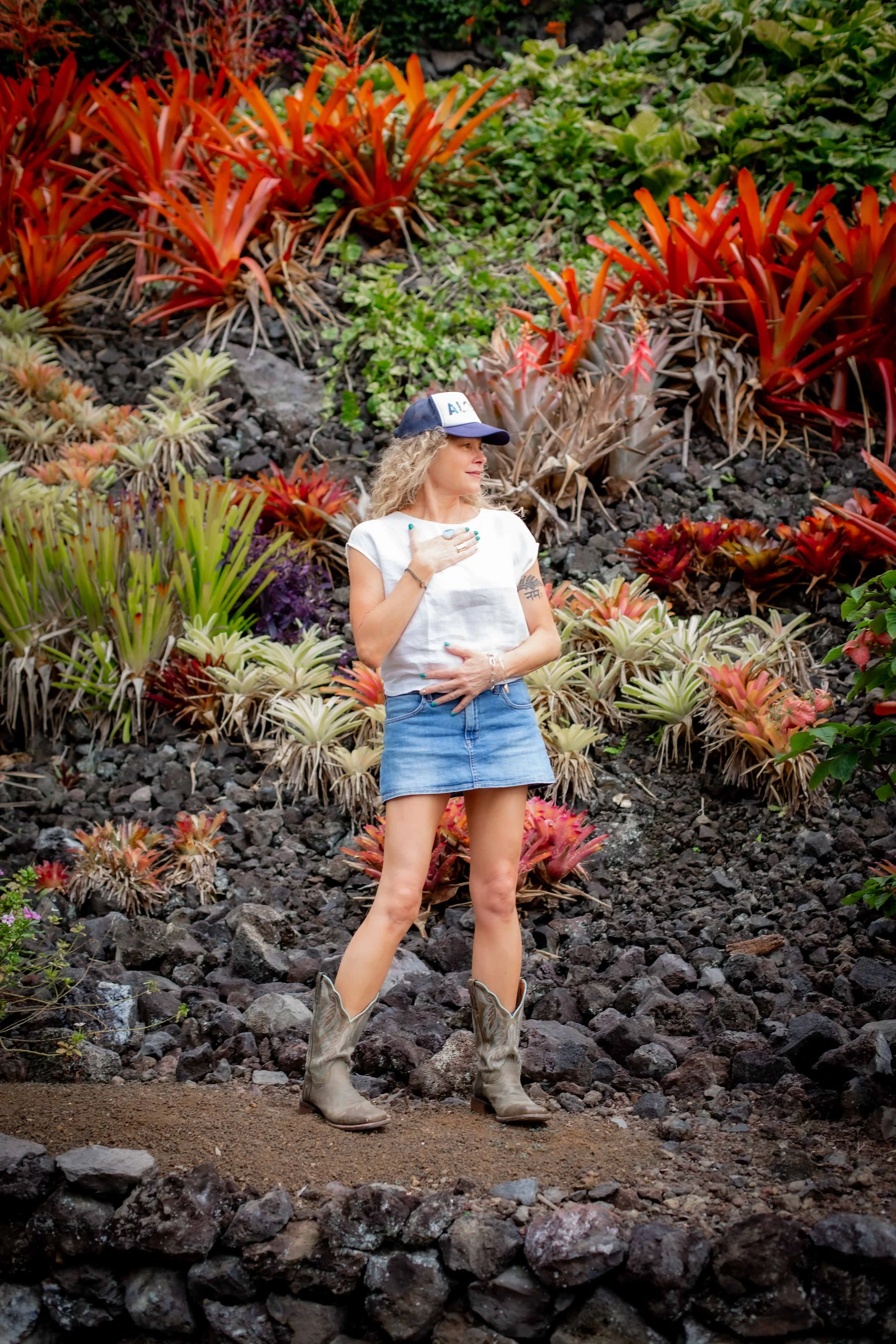 A woman with curly blonde hair, wearing a white tank top, a denim skirt, cowboy boots, and a baseball cap, standing on rocky terrain in front of a colorful garden with various succulents.