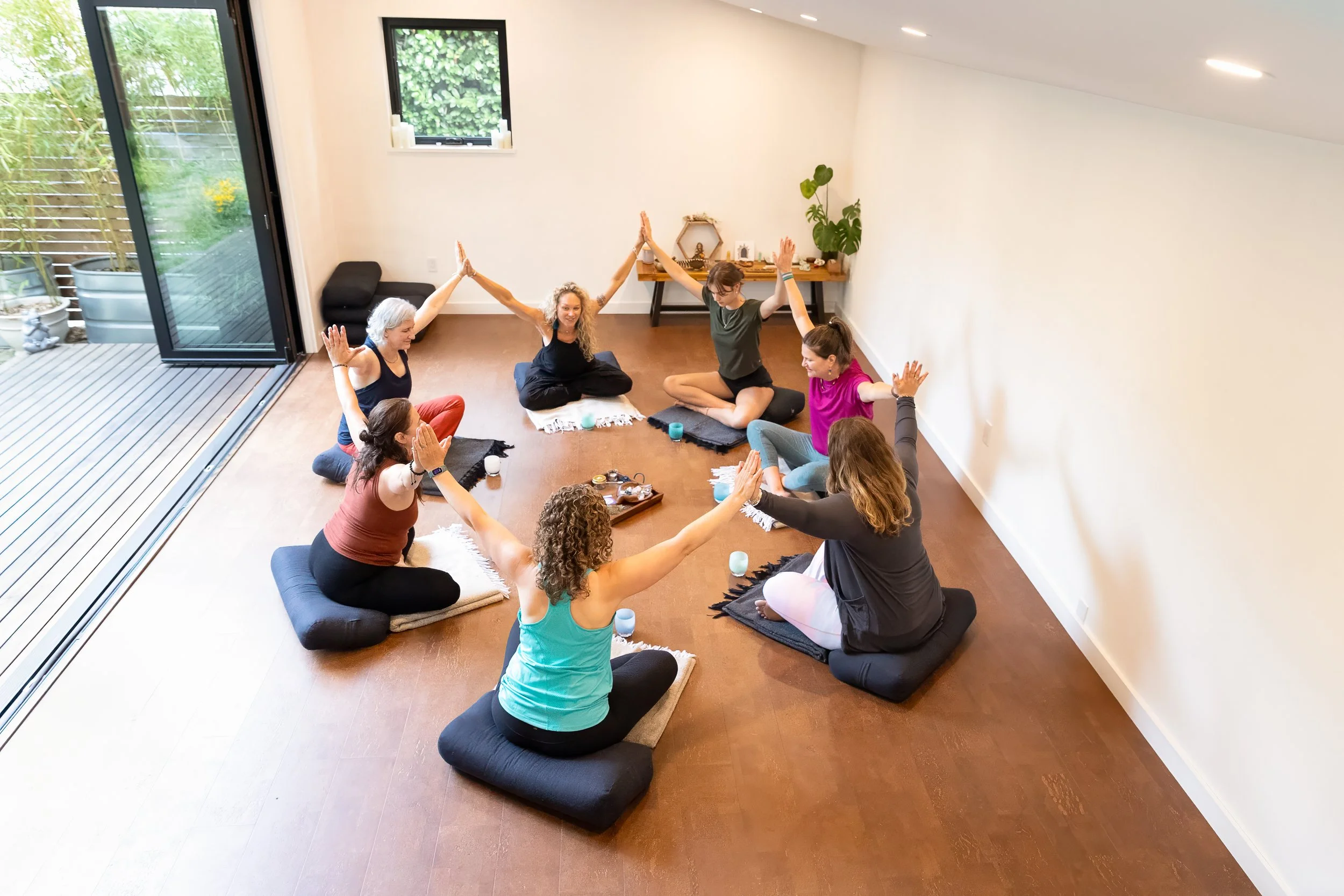 A group of people practicing yoga indoors on mats, sitting in a circle with hands raised and palms pressed together. Large windows show a patio and garden outside.