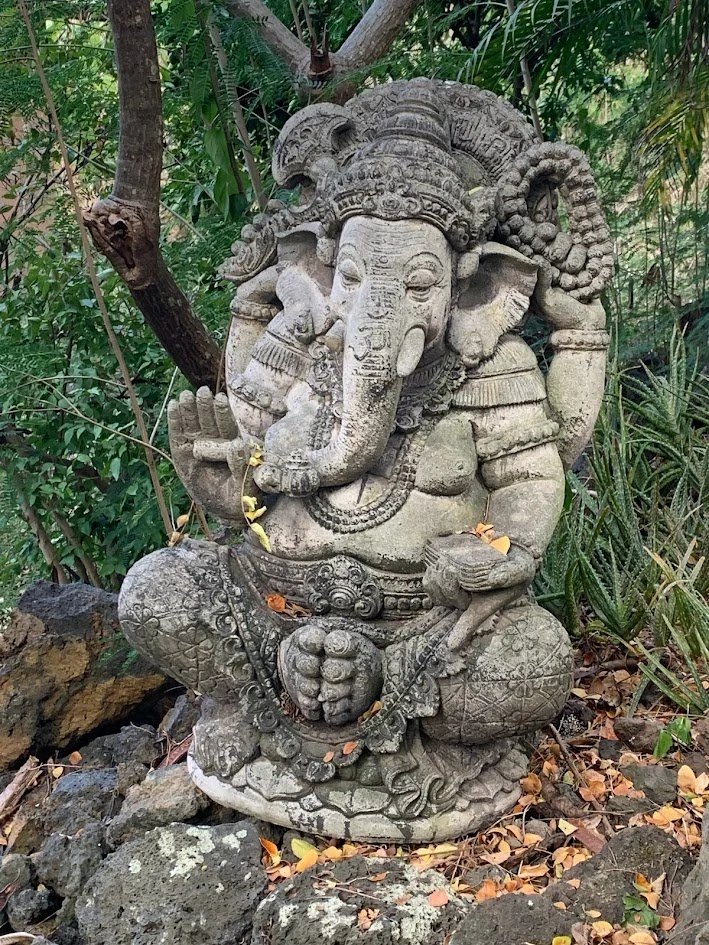 Stone sculpture of the Hindu god Ganesha sitting outdoors with greenery in the background.