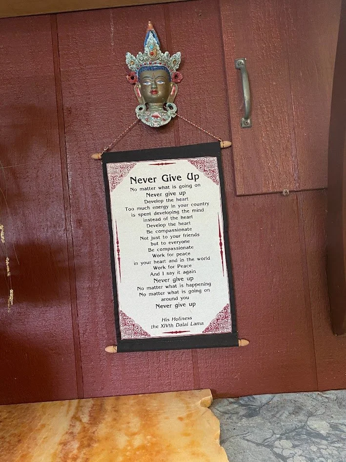 A decorative traditional mask or face sculpture hanging above a motivational poster on a wooden wall. The poster features a poem titled 'Never Give Up' by His Holiness the XIVth Dalai Lama.