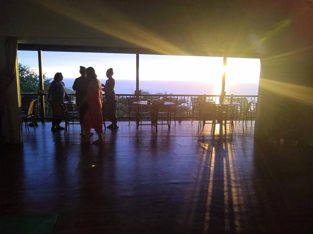 Interior of a room with a large open sliding door, overlooking a balcony with a view of the ocean at sunset. Four people are silhouetted against the bright sunset, standing and talking, with chairs and tables on the balcony.