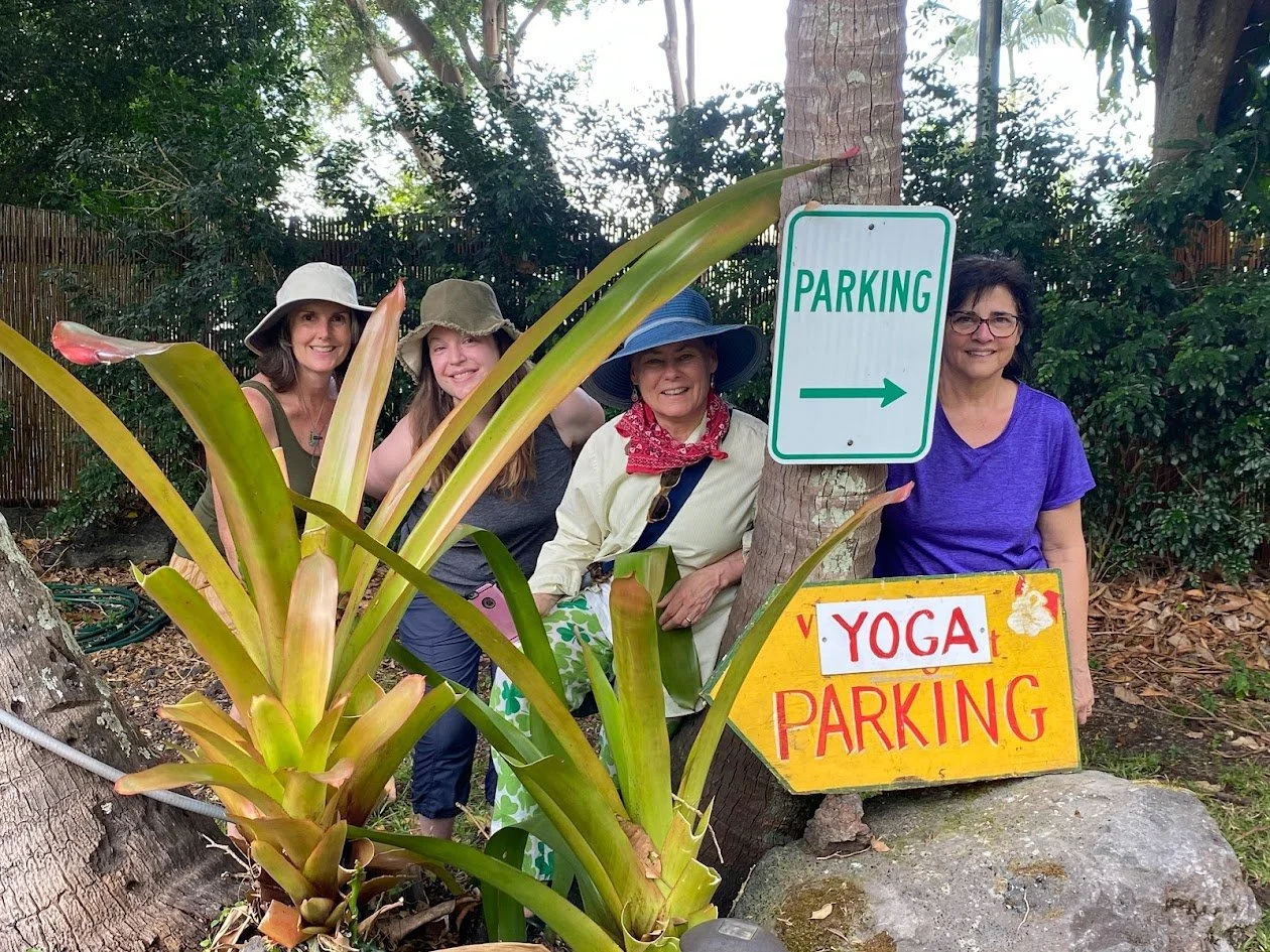 Four women outdoors standing behind large tropical plants, with signs reading 'Parking' and 'Yoga Parking,' surrounded by trees and greenery.