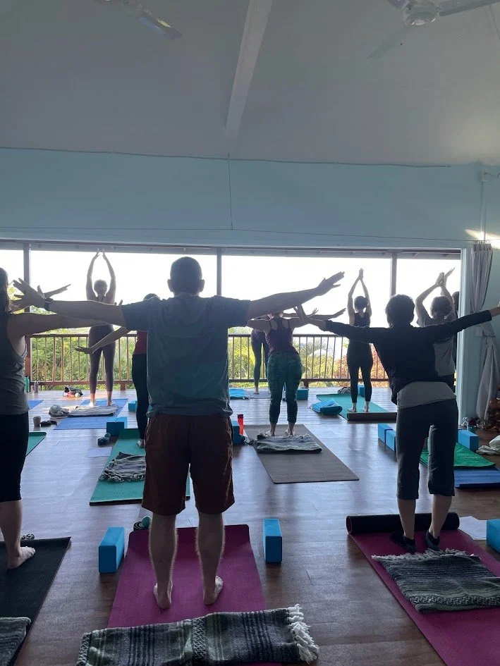 A group of people practicing yoga in a brightly lit studio with large windows, doing standing poses with arms outstretched.