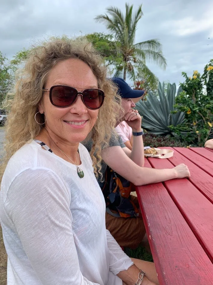 A woman with curly blonde hair wearing sunglasses and a white top, smiling while sitting at a red outdoor picnic table. In the background, a man wearing a cap and a woman are sitting at the table with food, with tropical plants and a palm tree behind