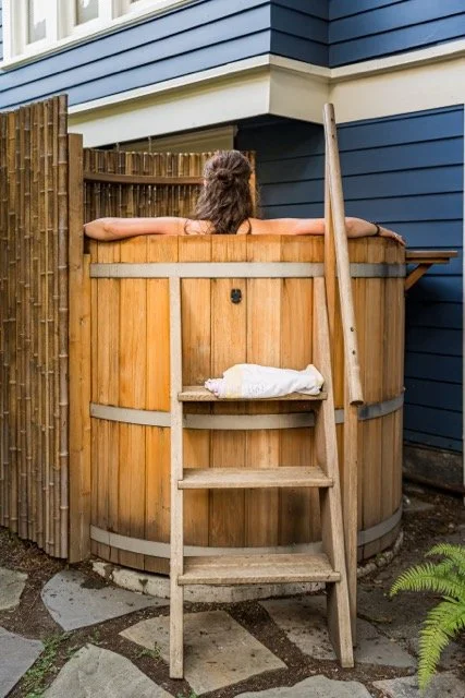 Person relaxing in a wooden hot tub in a backyard, with a ladder leaning against it and a towel on a shelf.