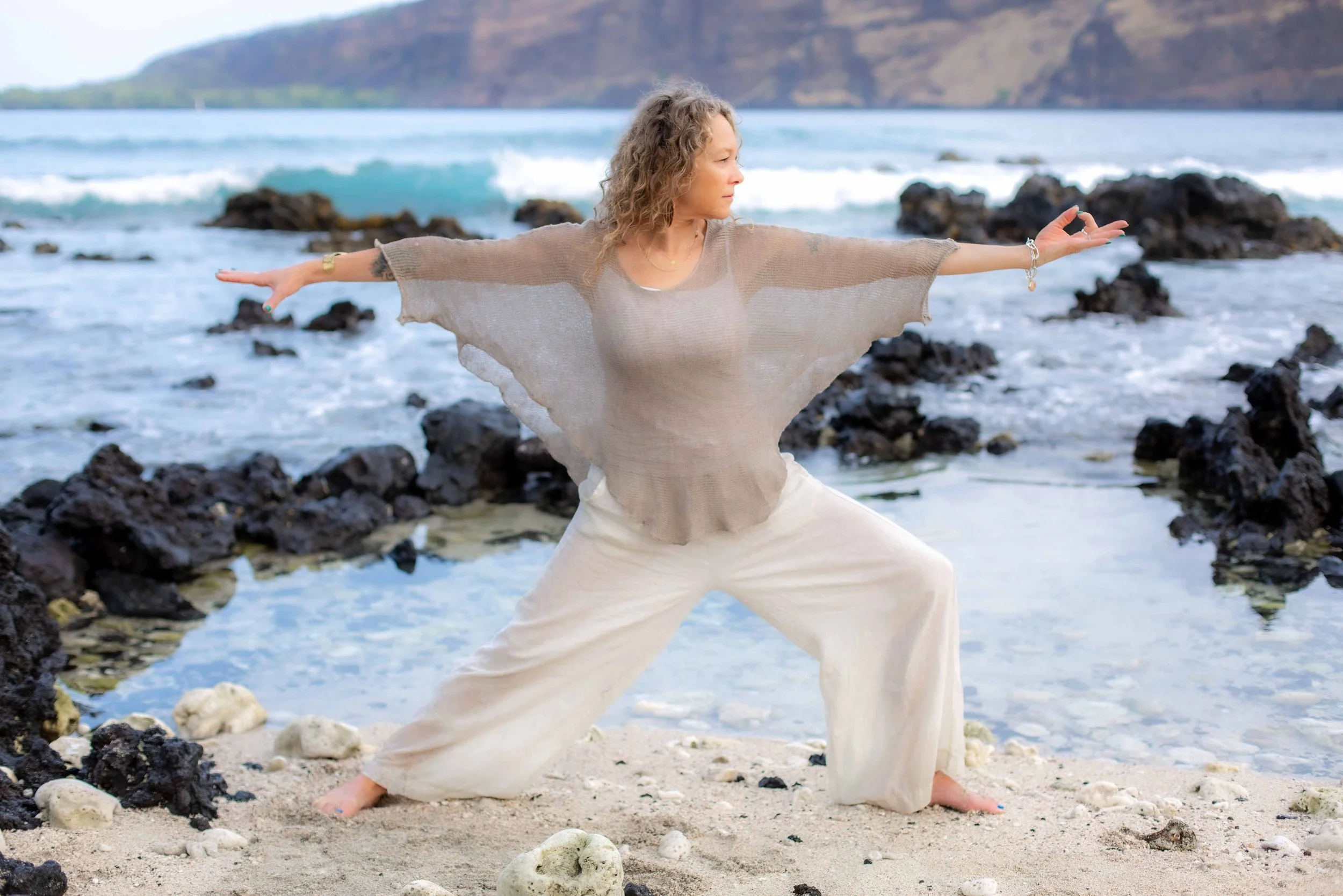 A woman with curly hair practicing yoga or dance on a rocky beach near the ocean, with mountains in the background.