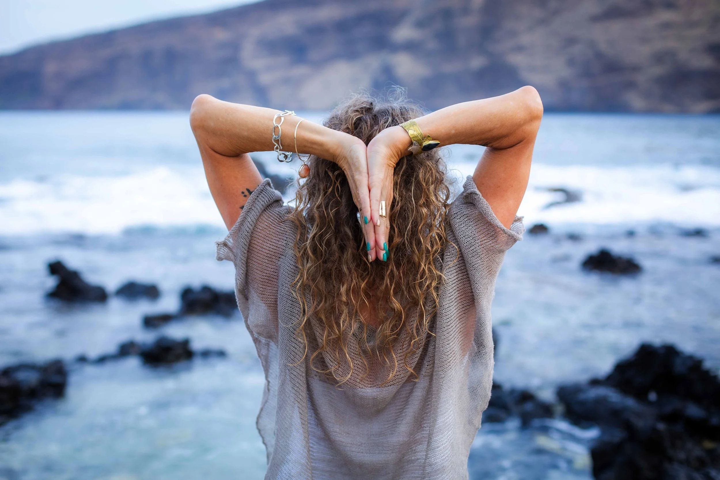 Person with long curly hair practicing yoga or stretching on a rocky beach near the water with mountains in the background.