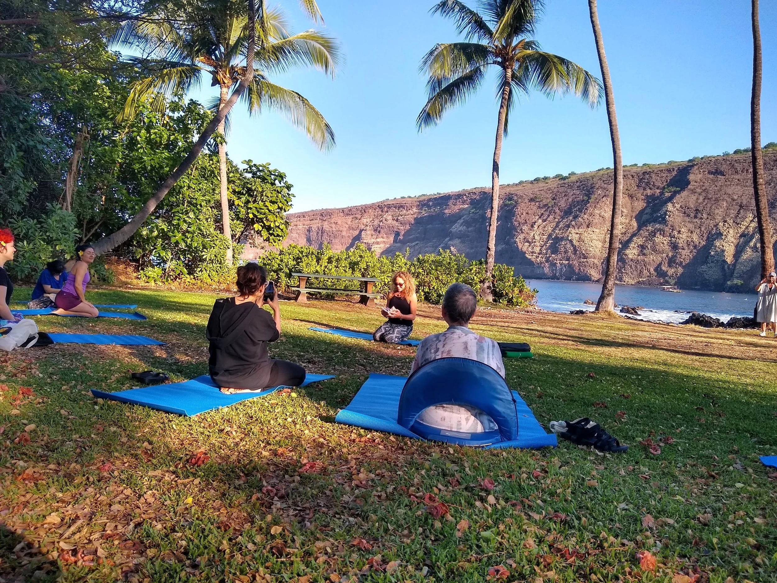 A group of people practicing yoga outdoors on mats near a beach, with palm trees, green bushes, and mountains in the background.