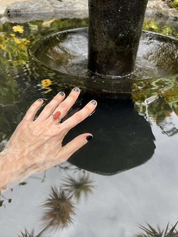 A hand with painted nails and a ring touching the water in a fountain or pond, with a tree trunk in the center and reflections of plants in the water.