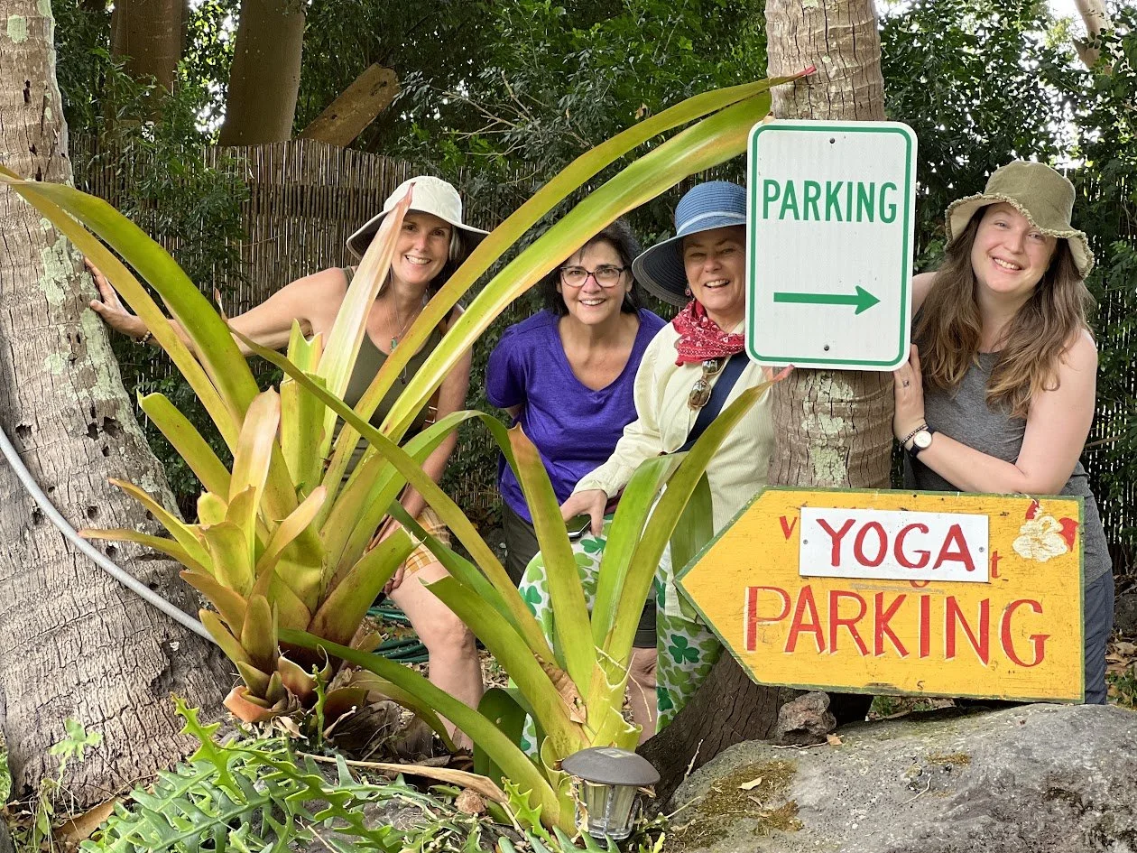 Four women standing behind a sign that says 'Yoga Parking' with an arrow pointing to the right, in a garden with large green plants and trees, some wearing sun hats, smiling at the camera.