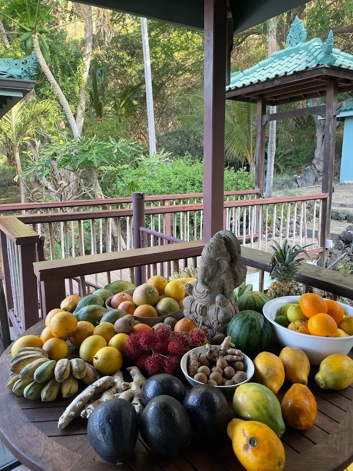 A table filled with various tropical fruits, a Buddha statue, and a pineapple, set outdoors with a wooden railing and lush greenery in the background.