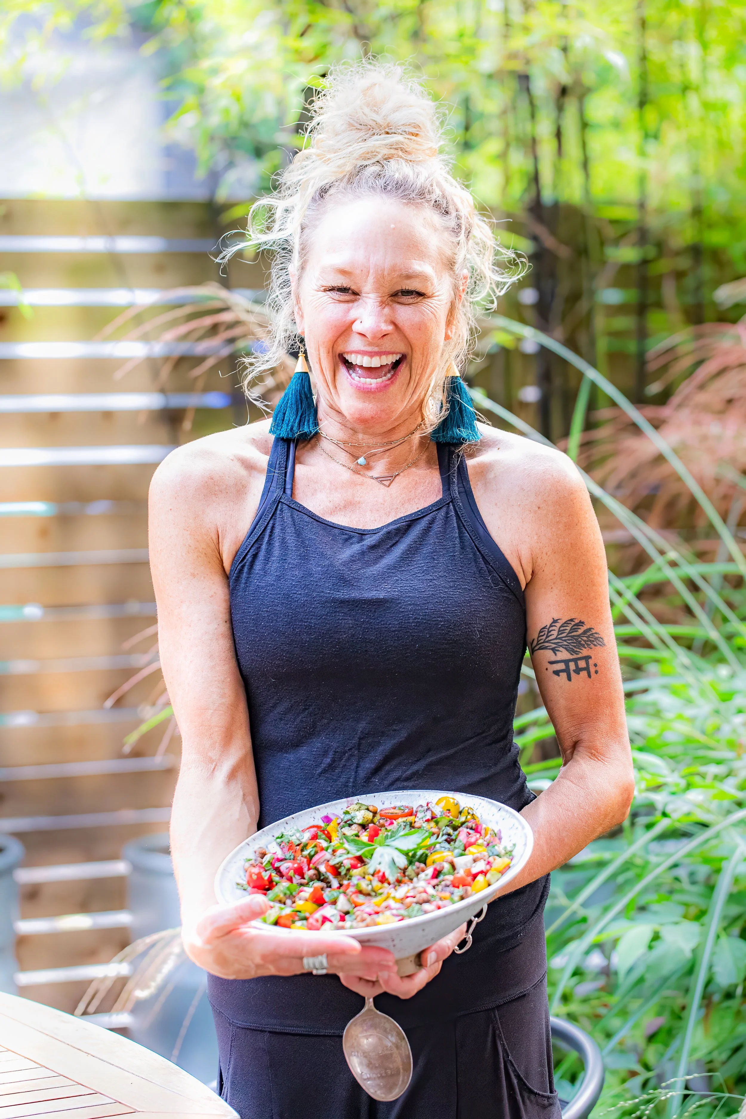 A woman with curly blonde hair in a bun, wearing a navy tank top and teal tassel earrings, smiles while holding a large bowl of colorful salad outdoors, with a wooden fence and greenery in the background.