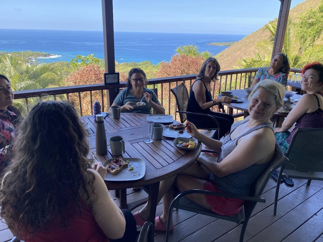 Group of women enjoying breakfast at a table on a balcony overlooking lush greenery, trees, hills, and the ocean in the background.