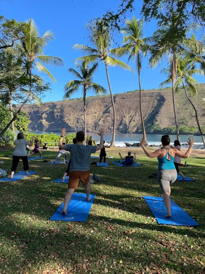 People participating in a yoga class outdoors near a lake, with palm trees and mountains in the background.