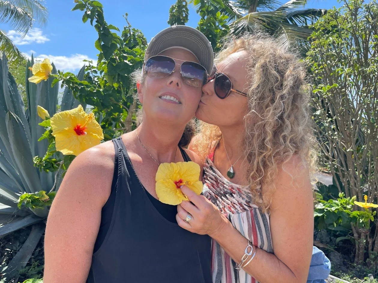 Two women in sunglasses sharing a kiss outdoors amidst tropical plants and yellow hibiscus flowers on a sunny day.