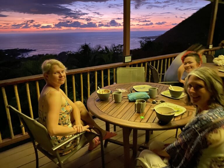 Three women sitting at a wooden outdoor table on a balcony overlooking the ocean during sunset, with bowls, cups, and napkins on the table.