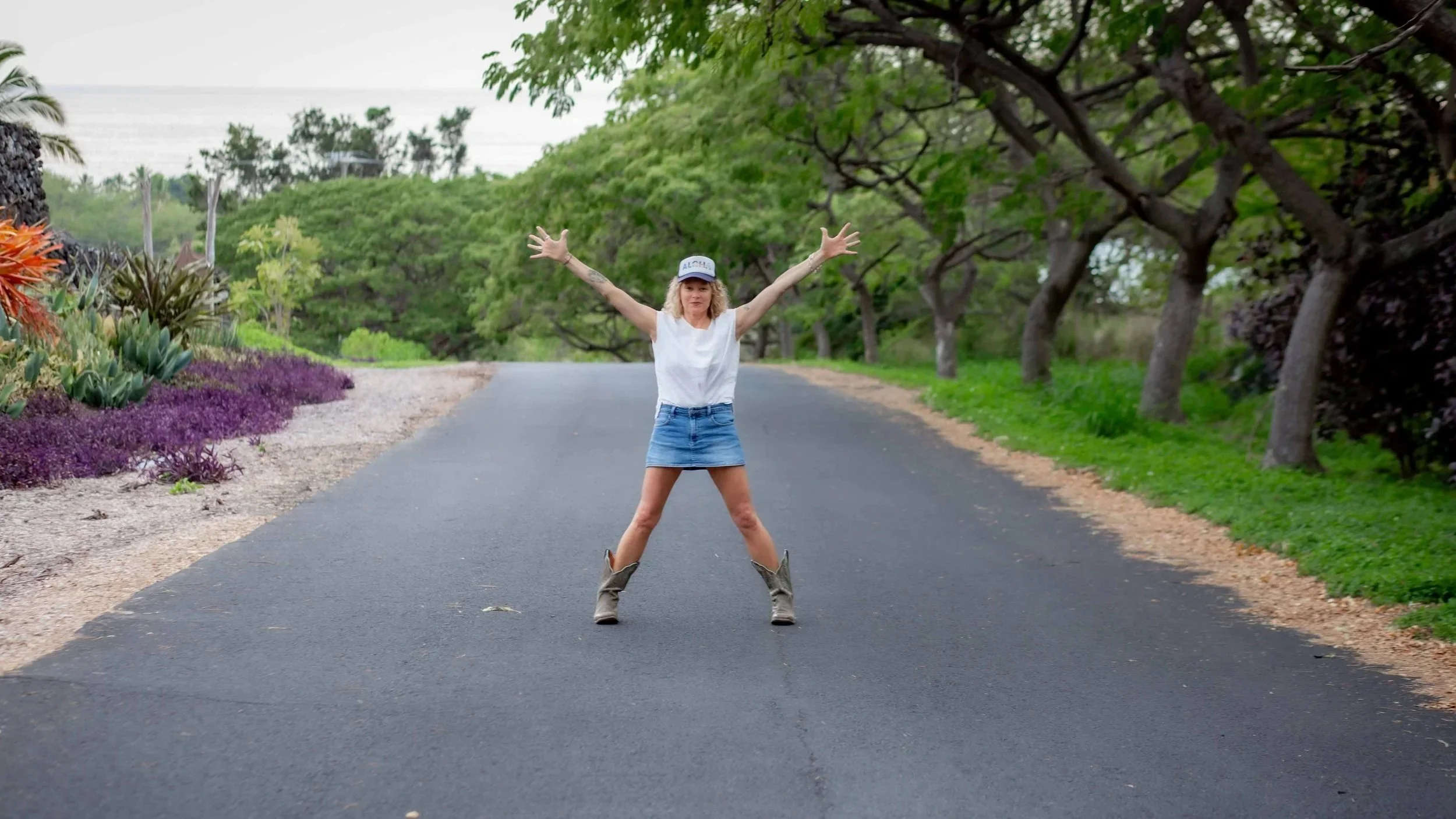 Woman in a white top and denim skirt wearing cowboy boots, standing with arms outstretched on a paved road surrounded by green trees and colorful plants.