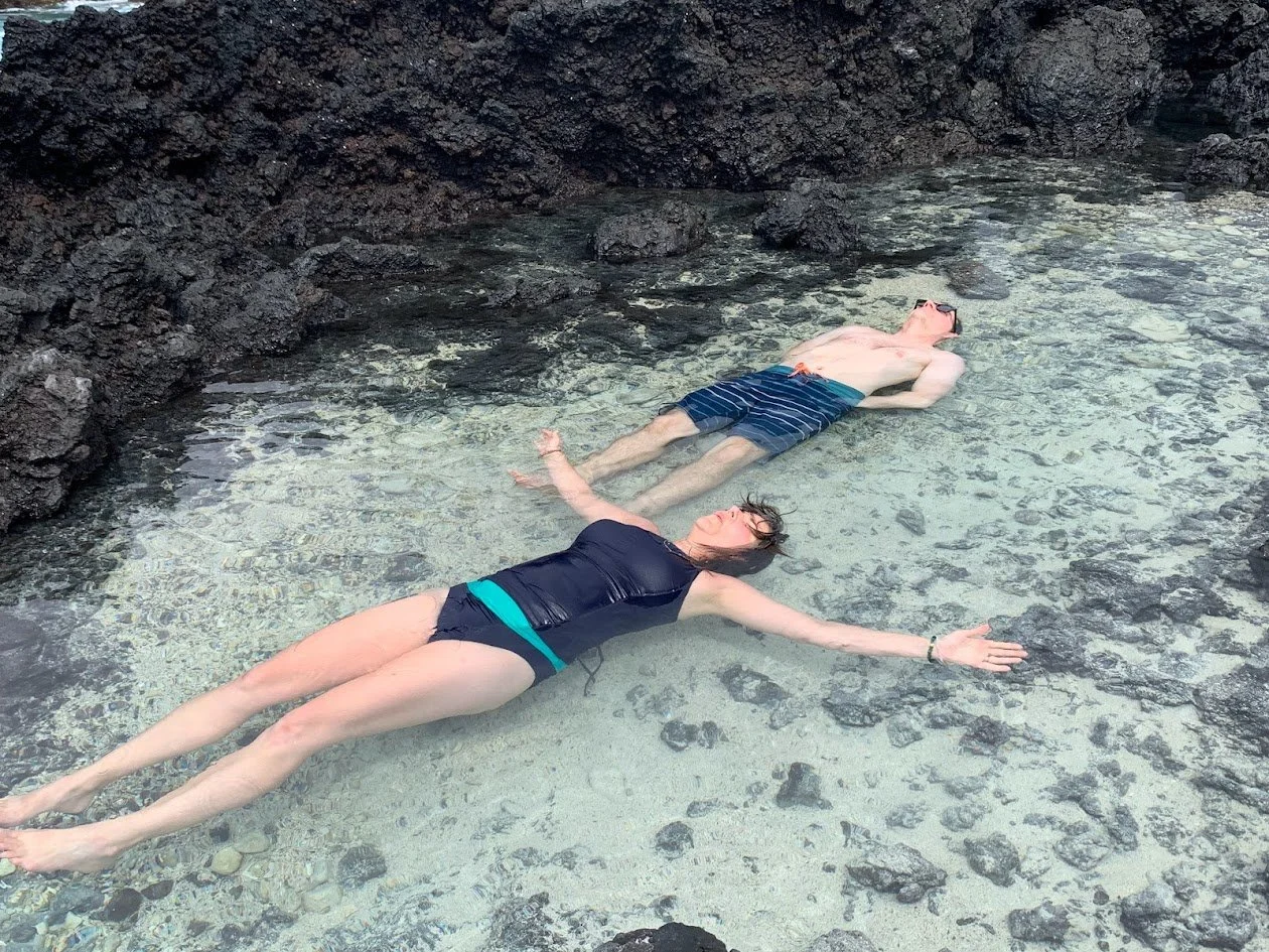 Two people, a man and a woman, are lying on their backs in shallow water near rocky terrain, relaxing with eyes closed and arms spread out.