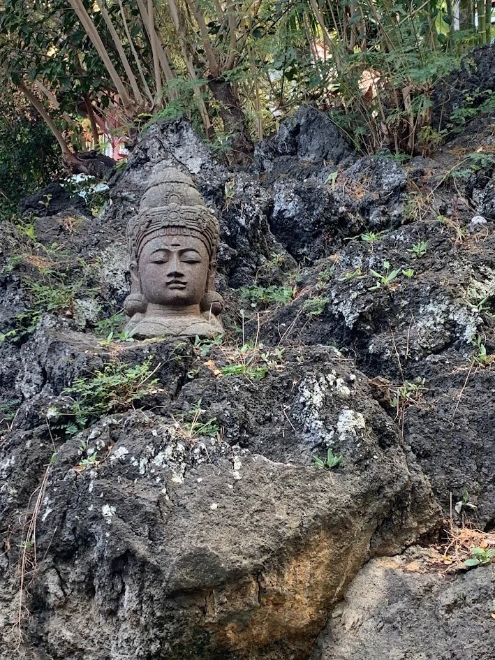 Stone sculpture of a serene face with detailed headwear, embedded in dark volcanic rocks with sparse greenery around.