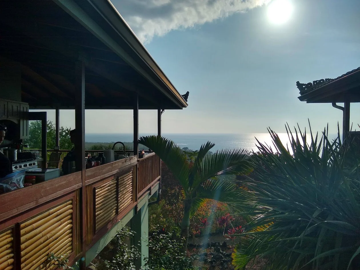 Balcony with wooden railing on a hillside overlooking the ocean, with tropical plants in the foreground and the sun shining in the sky.