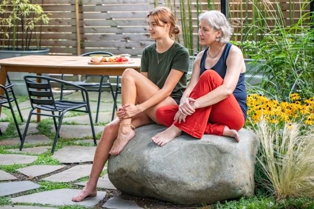 Two women sitting on a large rock in a backyard garden with greenery and yellow flowers, with a wooden fence and an outdoor table in the background.