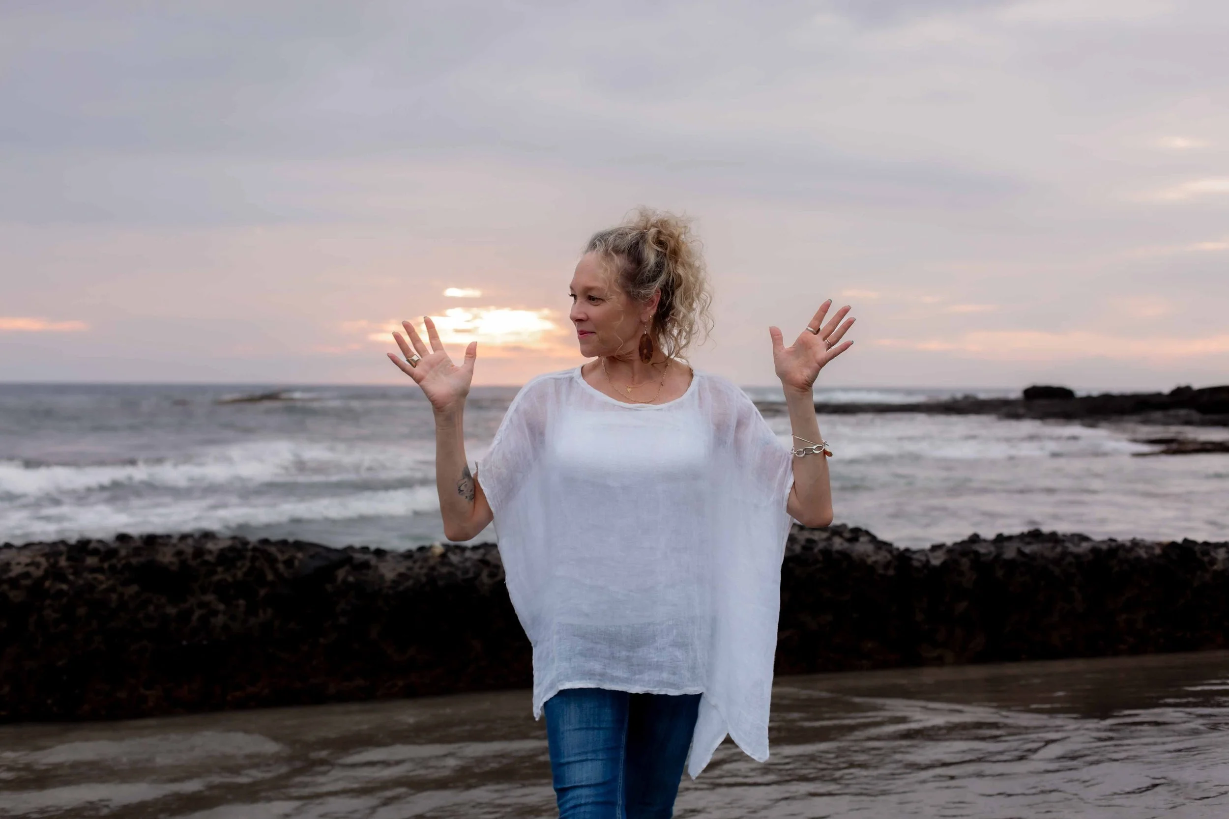 A woman with curly blonde hair standing on a beach at sunset, wearing a white top and blue jeans, with her arms raised and hands open.