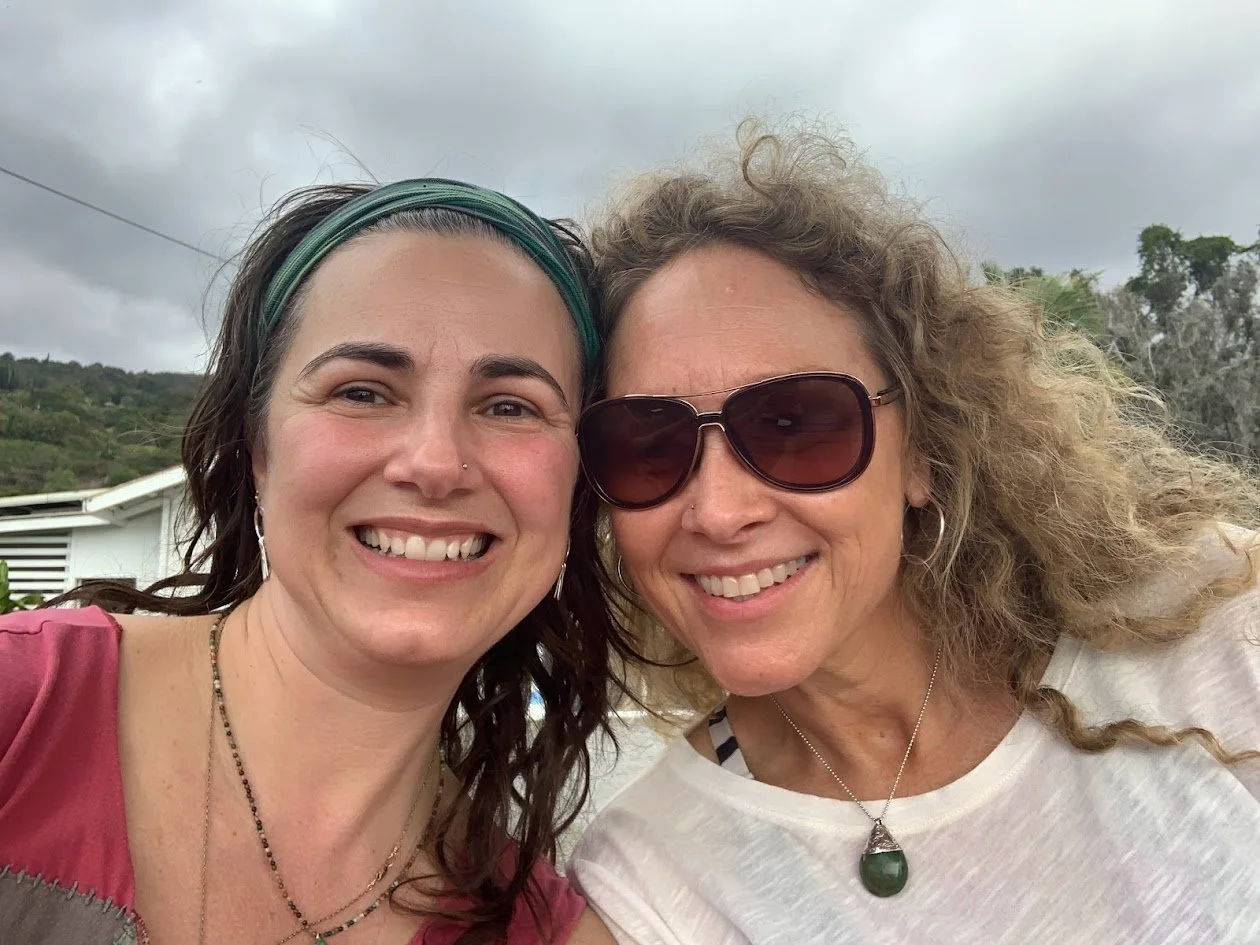 Two women smiling for a selfie outdoors, with cloudy sky and greenery in the background.