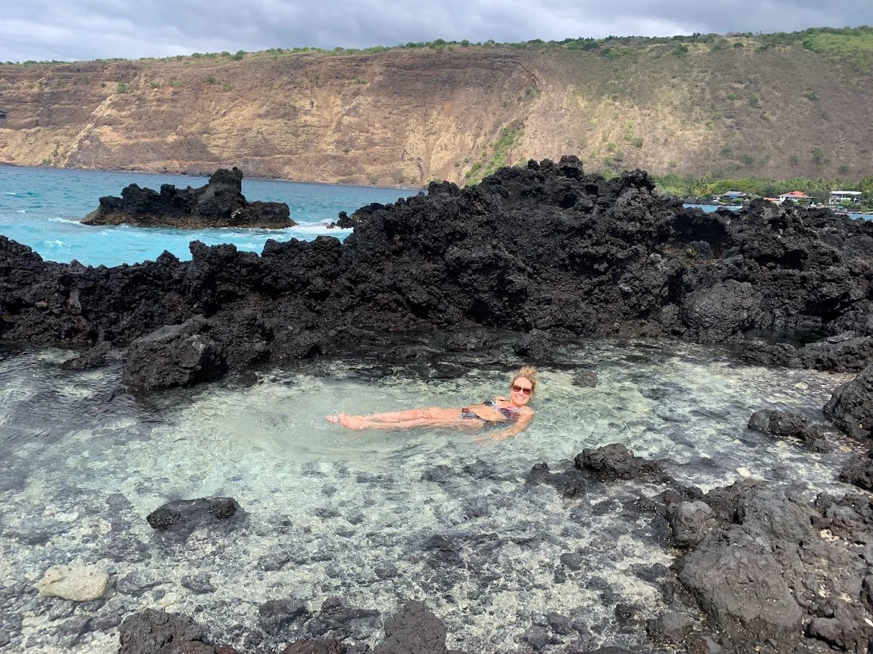 A woman in sunglasses relaxing in a small, natural tide pool surrounded by black volcanic rocks with a scenic ocean and cliffside in the background.