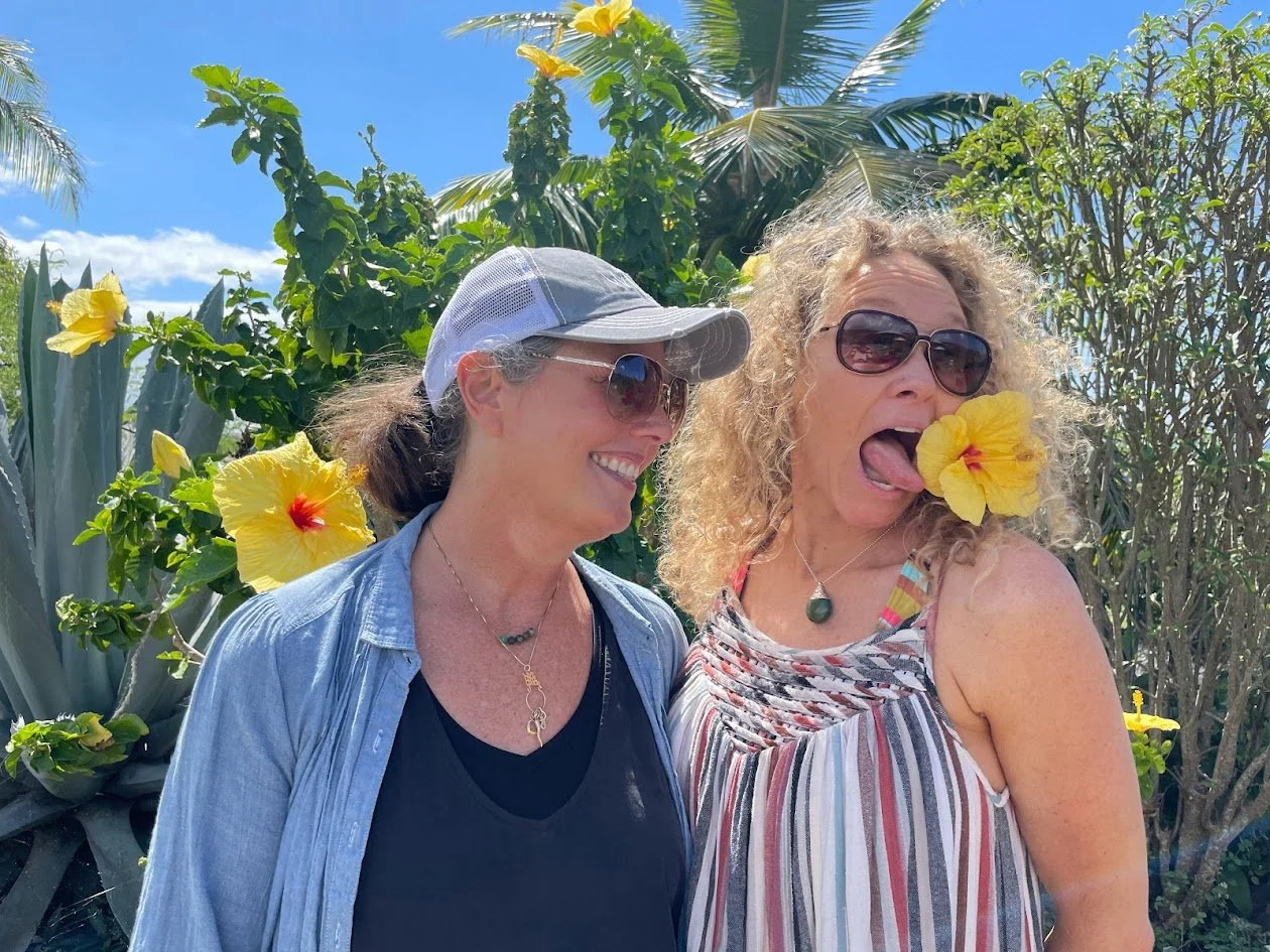 Two women enjoying a day outdoors in a garden with yellow hibiscus flowers; one woman is smiling and wearing sunglasses and a baseball cap, the other is sticking out her tongue with a hibiscus flower in her mouth, wearing sunglasses and a striped dre