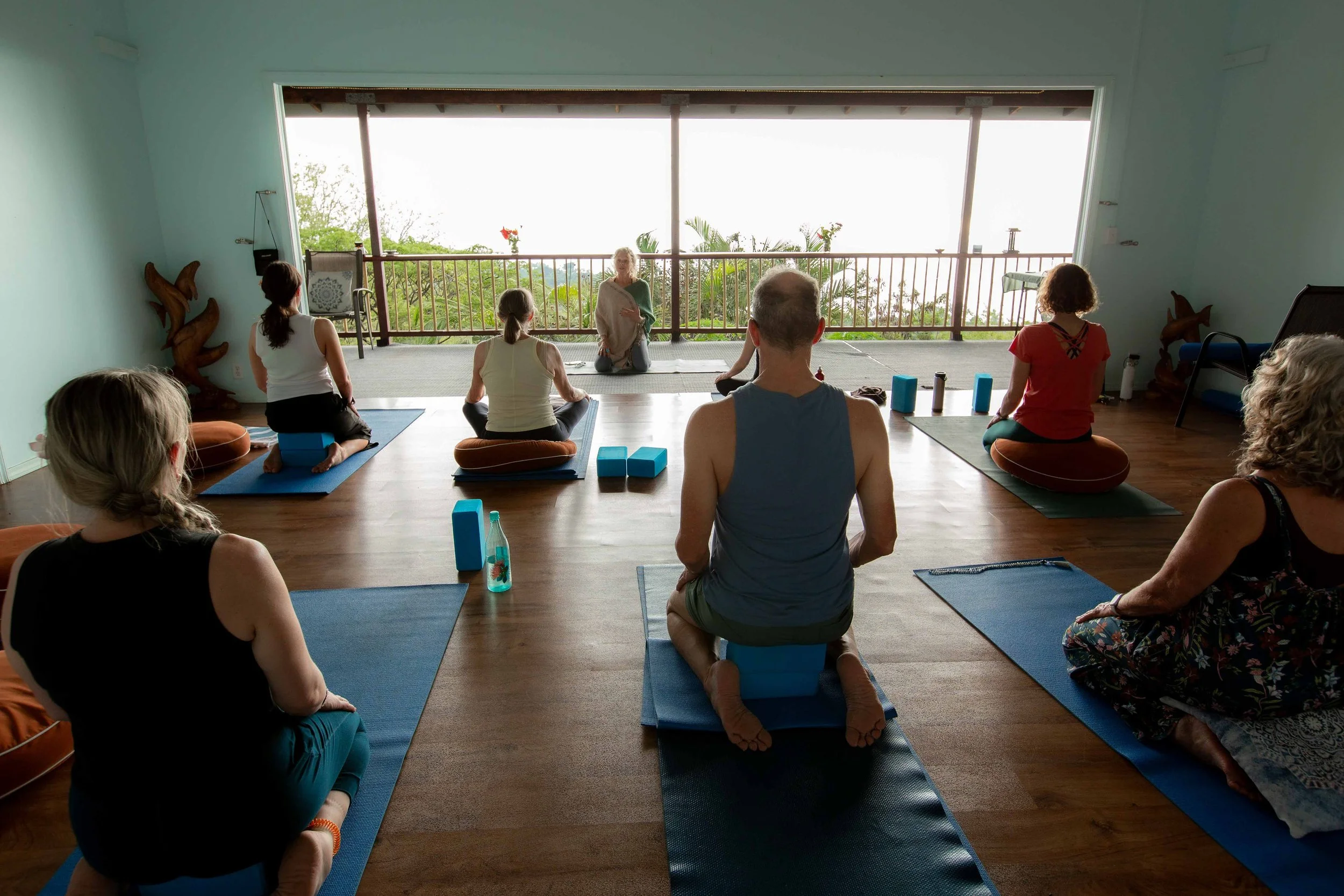 Participants in a yoga or meditation class seated on mats in a room with large open windows overlooking greenery; instructor at front leading the session.