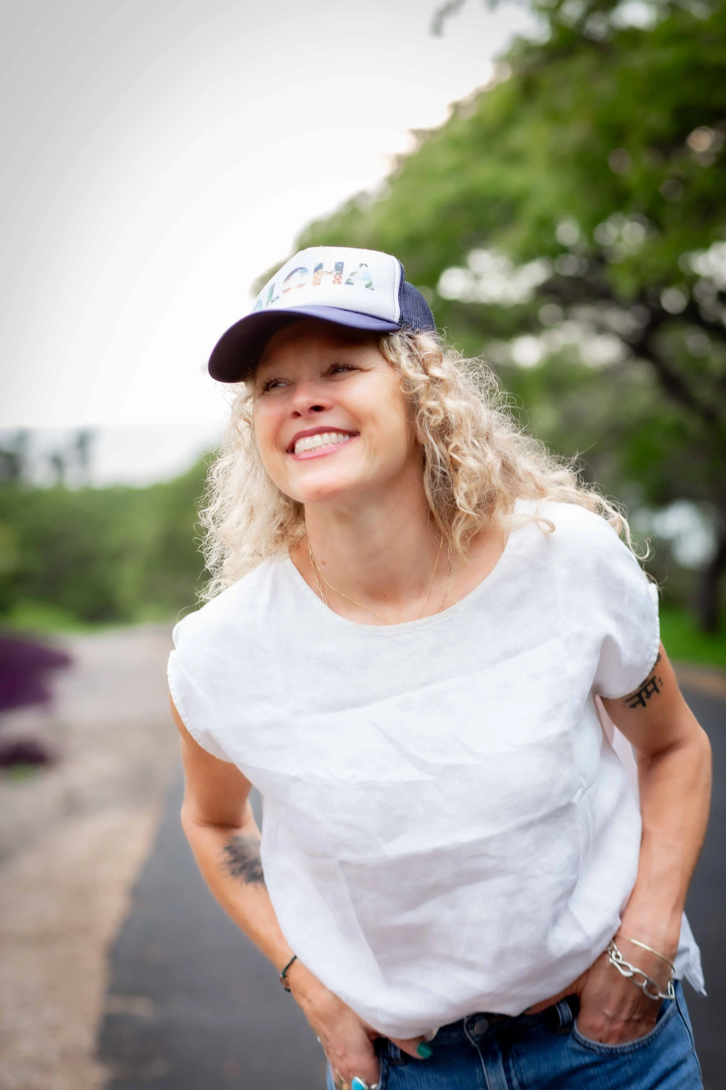A smiling woman with curly blonde hair wearing a white t-shirt and a baseball cap with the word 'ALOHA' on it, standing outdoors on a paved path with greenery in the background.
