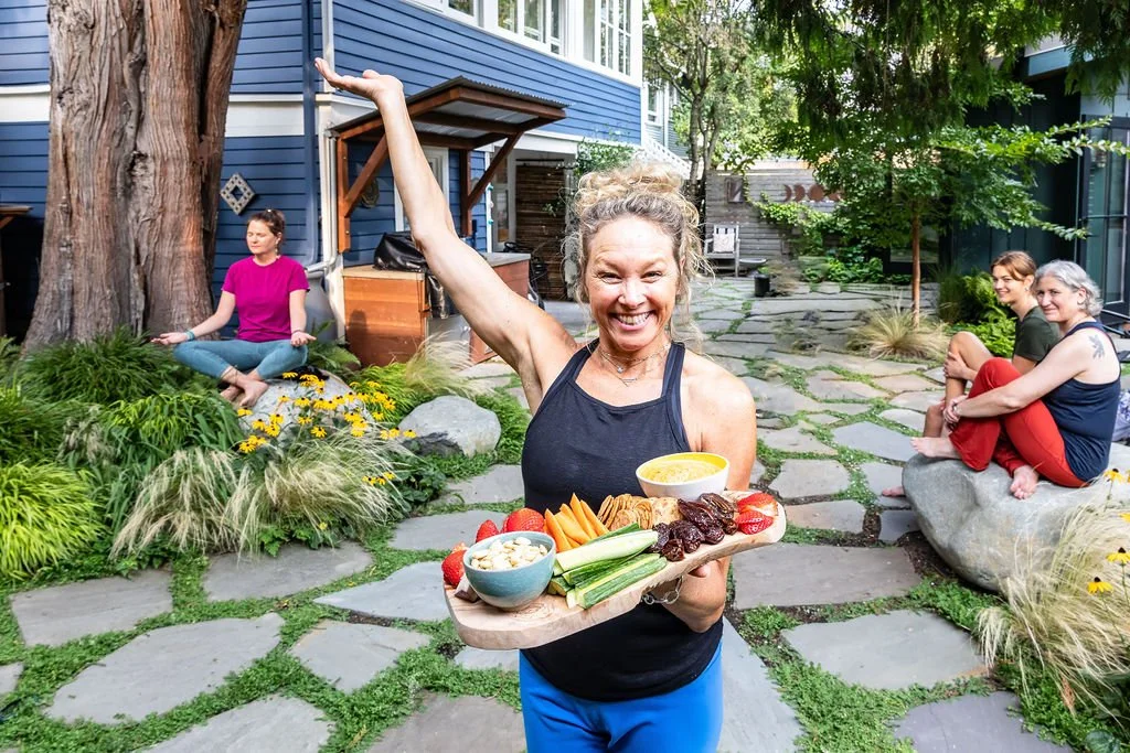 A woman in athletic wear smiling and holding a tray of healthy food, including fruit, vegetables, and dip, outdoors on a stone pathway with three other women sitting on rocks and a bench nearby, surrounded by greenery and a blue house in the background.