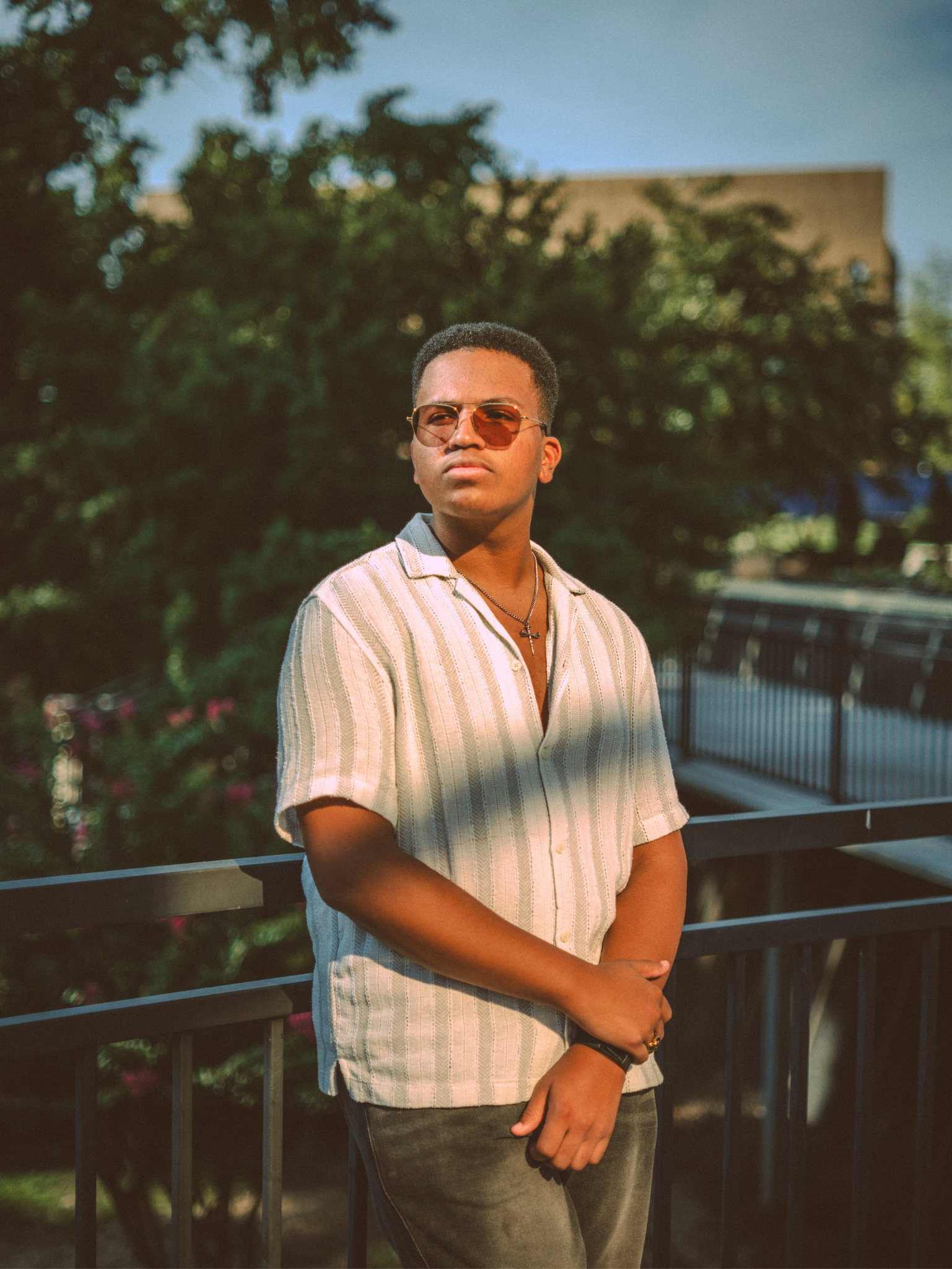 A young man with short hair wearing sunglasses, a light-colored striped short-sleeve shirt, dark jeans, and a cross necklace, standing outdoors on a balcony with a metal railing, in front of trees and a building, during sunset.