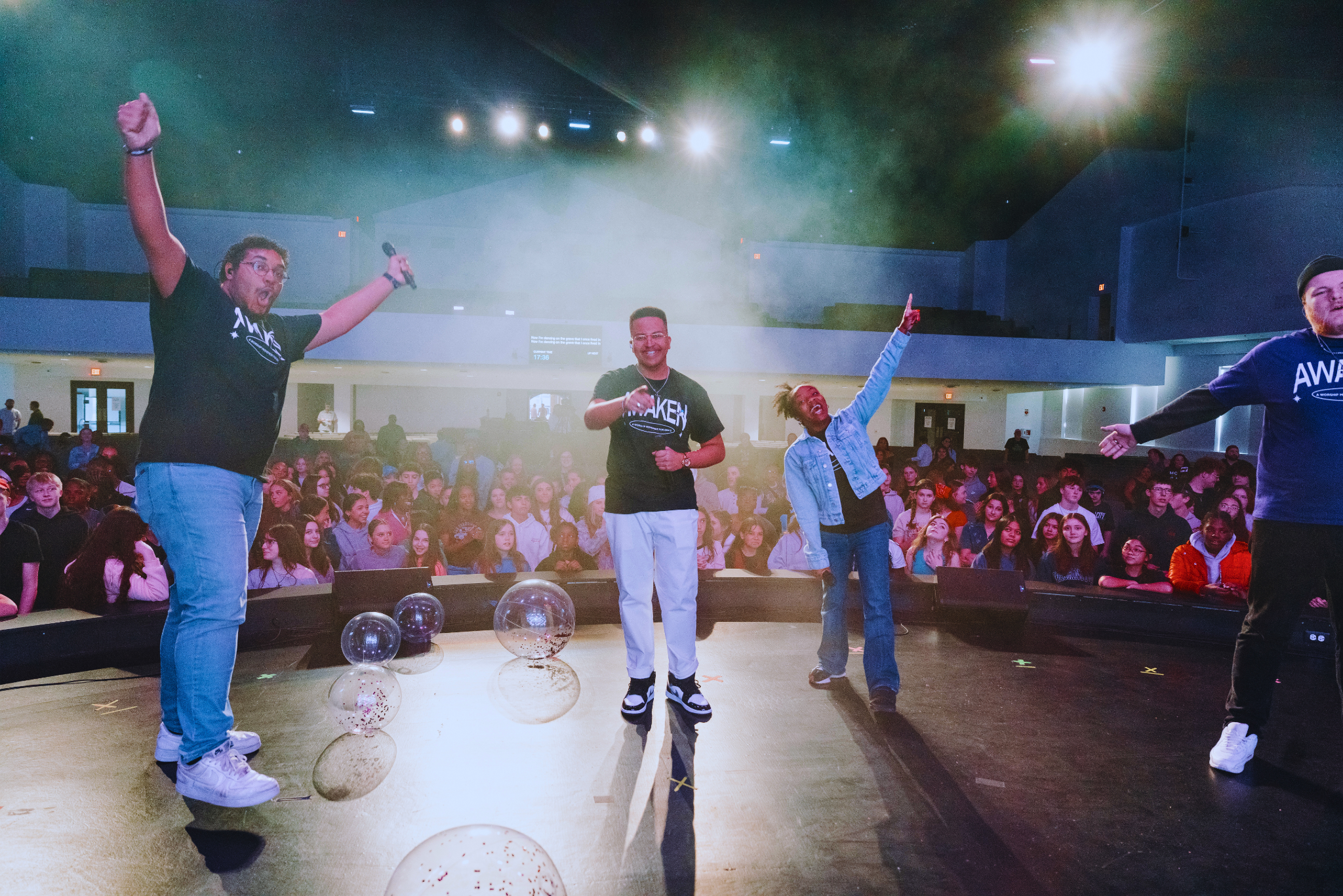 Four speakers on stage with microphone, leading a youth event in an auditorium filled with a seated mostly teenage audience, some balloons on the stage.