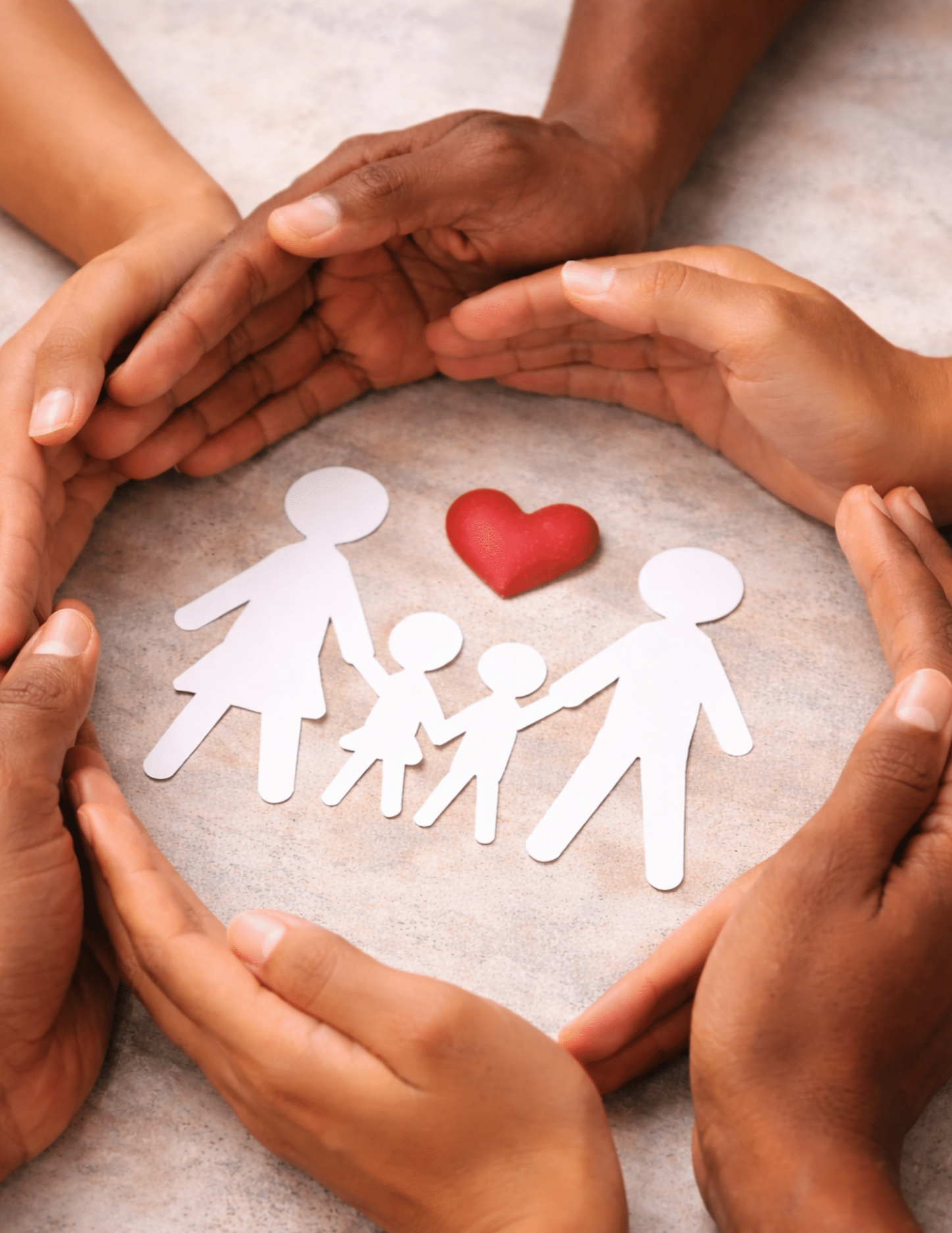 Multiple hands surrounding paper cutouts of a family, with a small red heart and a red heart-shaped object on a beige surface.