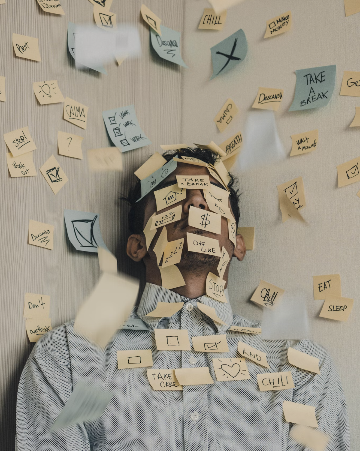 A man with sticky notes covering his face and body, with additional sticky notes scattered around him on a wall and floating in the air. The notes have various handwritten words and symbols like 'Take a break', 'Chill', 'Sleep', and checkmarks.