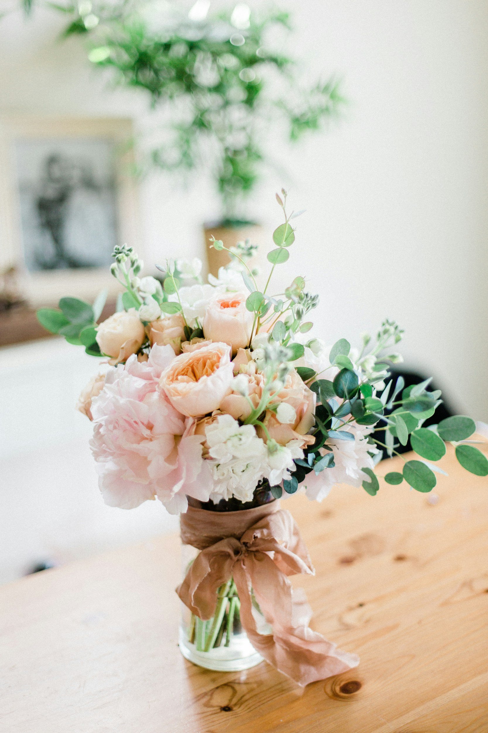 A bouquet of soft pink and white flowers with green foliage, tied with a tan ribbon, sitting in a clear glass jar on a wooden table.