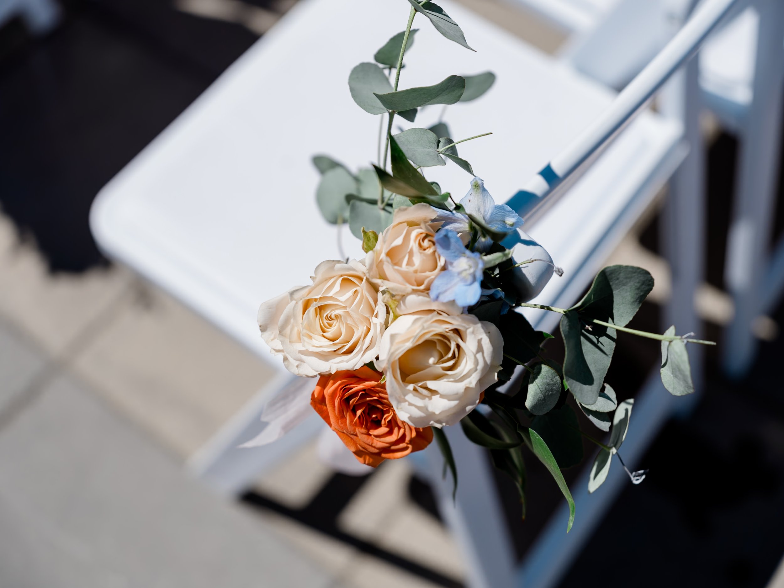 A small bouquet of cream, orange, and light blue flowers with green leaves resting on a white chair.