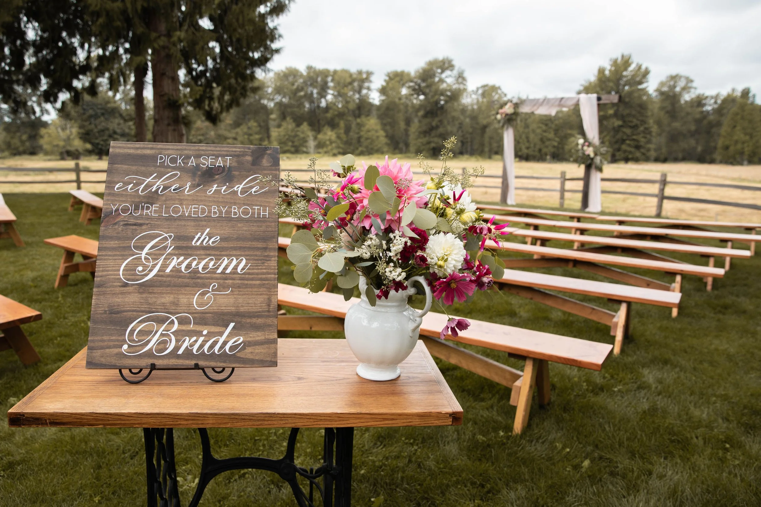 Wedding outdoor ceremony setup with a wooden sign on a table next to a vase of pink, white, and purple flowers, and a wooden arch decorated with white fabric and flowers in the background.