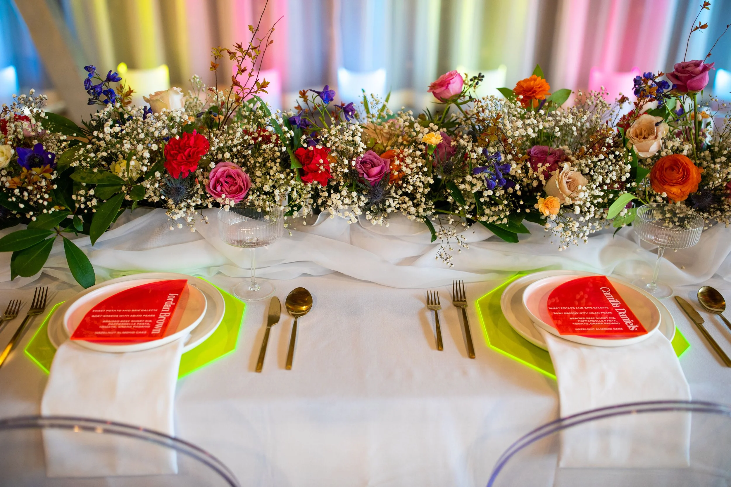 A beautifully set dining table with white plates, silver utensils, and transparent glasses. The table is decorated with a large, colorful floral centerpiece featuring roses, baby's breath, and other flowers. The background has colorful drapes, creati